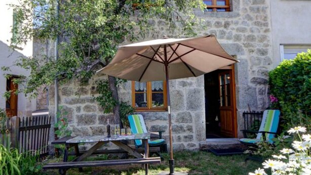 A stone building with a wooden door, a window, and a patio area with a table, chairs, and an umbrella. - Charming Cottage with Garden in Estivareilles (Estivareilles)