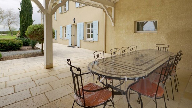 Een overdekt terras met een ronde tafel en stoelen, een pergola en een gebouw met lichtblauwe luiken op de achtergrond.