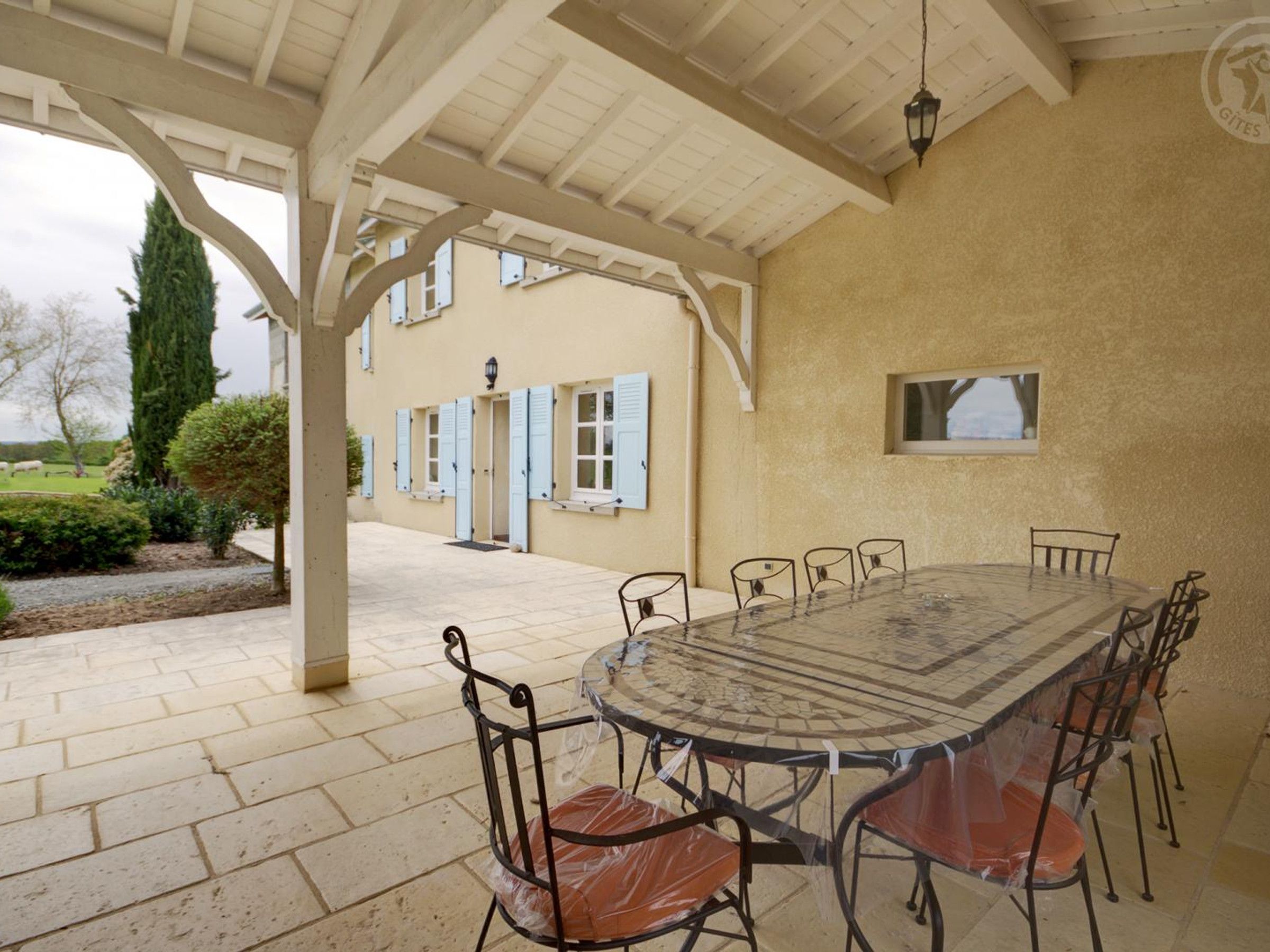 Zona pranzo all'aperto riparata, con tavolo e sedie rotonde, una struttura a pergola e un edificio con persiane blu sullo sfondo.