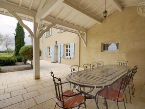A covered outdoor dining area with a round table and chairs, a pergola structure, and a building with blue shutters in the background. - Spacious house with heated pool, 4 bedrooms in Chalain-le-Comtal (Chalain-le-Comtal)