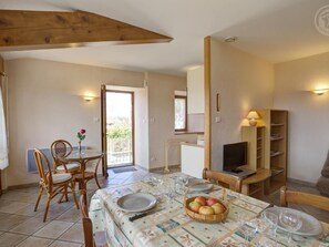 A dining area with a table set for four, surrounded by wicker chairs. A television is mounted on the wall, and a bookshelf is visible to the right.