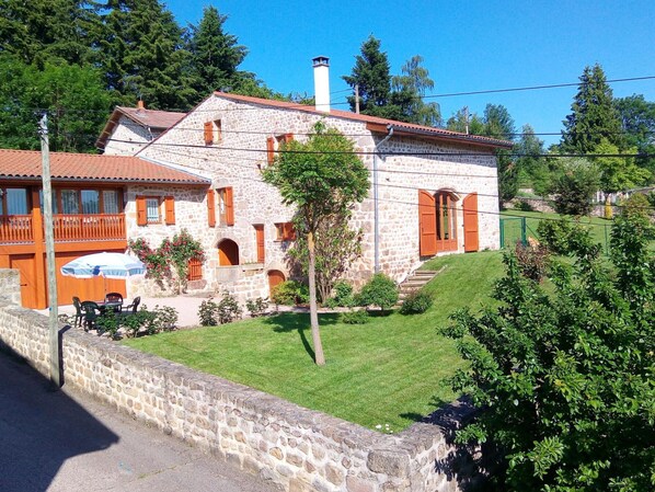 A stone building with a red-tiled roof, a balcony with wooden railings, and a small table with chairs outside.