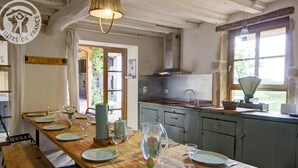 A rustic dining area with a long wooden table set for a meal, a kitchen with a stove and cabinets, and a view of greenery outside.