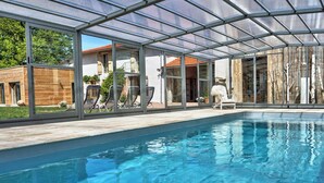 A modern indoor swimming pool with glass walls, a wooden deck, and lounge chairs.