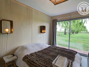 Une chambre avec un grand lit, une table de chevet, une lampe et une vue sur un espace verdoyant par la fenêtre.
