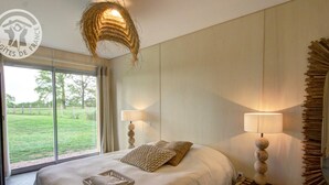 A bedroom with a large bed, two bedside lamps, a wicker ceiling light, and a view of a green landscape through a sliding door.
