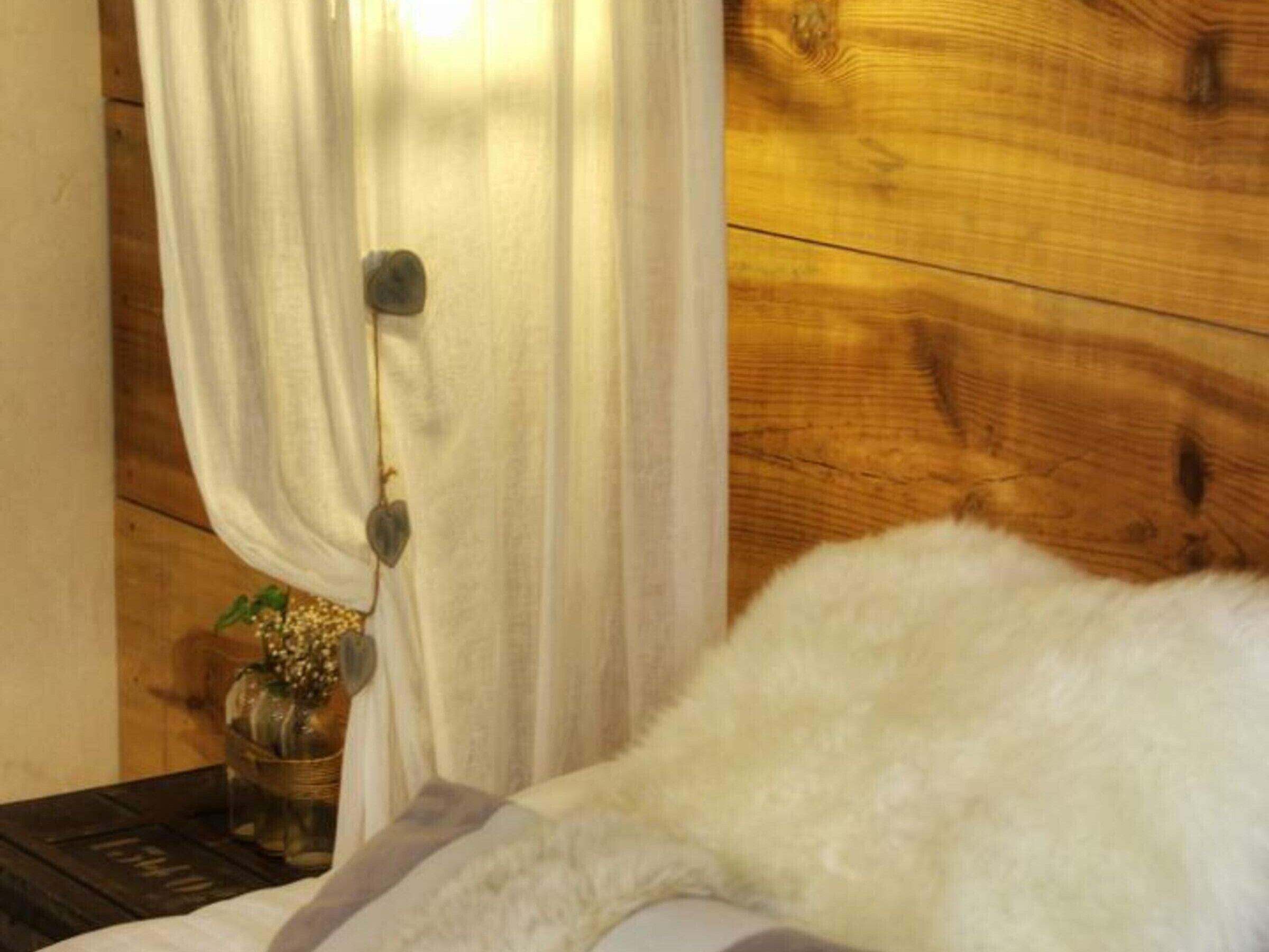 A room with a wooden wall, a white curtain, a small table with a vase of flowers, and a fluffy white pillow on a bed.