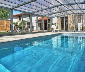 Una piscina interior moderna con paredes de vidrio, una terraza de madera y sillas de descanso.