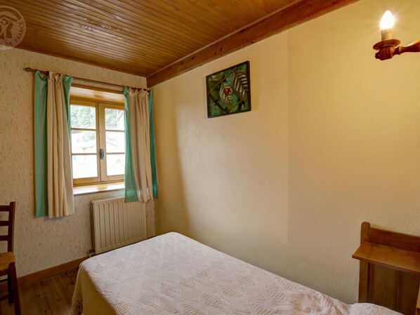 Una habitación con techo de madera, una cama con colcha blanca estampada, un radiador, una ventana con cortinas y una lámpara fijada a la pared.