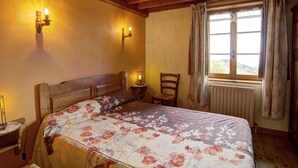 Une chambre confortable avec un plafond en bois, un grand lit avec une literie à motifs floraux, deux lampes fixées au mur, une fenêtre avec des rideaux et un radiateur.
