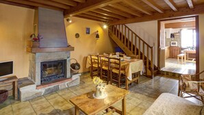 Une pièce chaleureuse avec une cheminée, des poutres apparentes en bois au plafond, un coin repas avec une table et des chaises, et une cuisine visible à travers une porte ouverte.