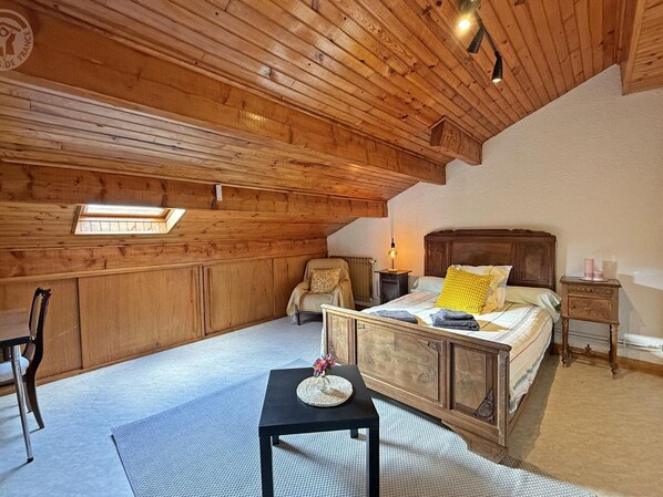 A cozy bedroom with a wooden ceiling, a bed with a wooden headboard, a small table with a mat, and a chair.