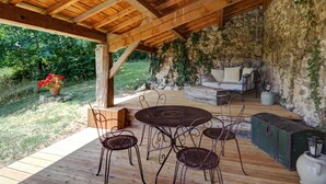 A covered outdoor seating area with a stone wall, wooden ceiling, and a view of greenery. - Authentic cottage for 12 with terrace in countryside (Saint-Just-la-Pendue)