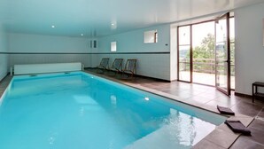 A large indoor swimming pool with clear blue water, surrounded by lounge chairs and a glass door leading to a balcony with a view of greenery. - Authentic cottage for 12 with terrace in countryside (Saint-Just-la-Pendue)