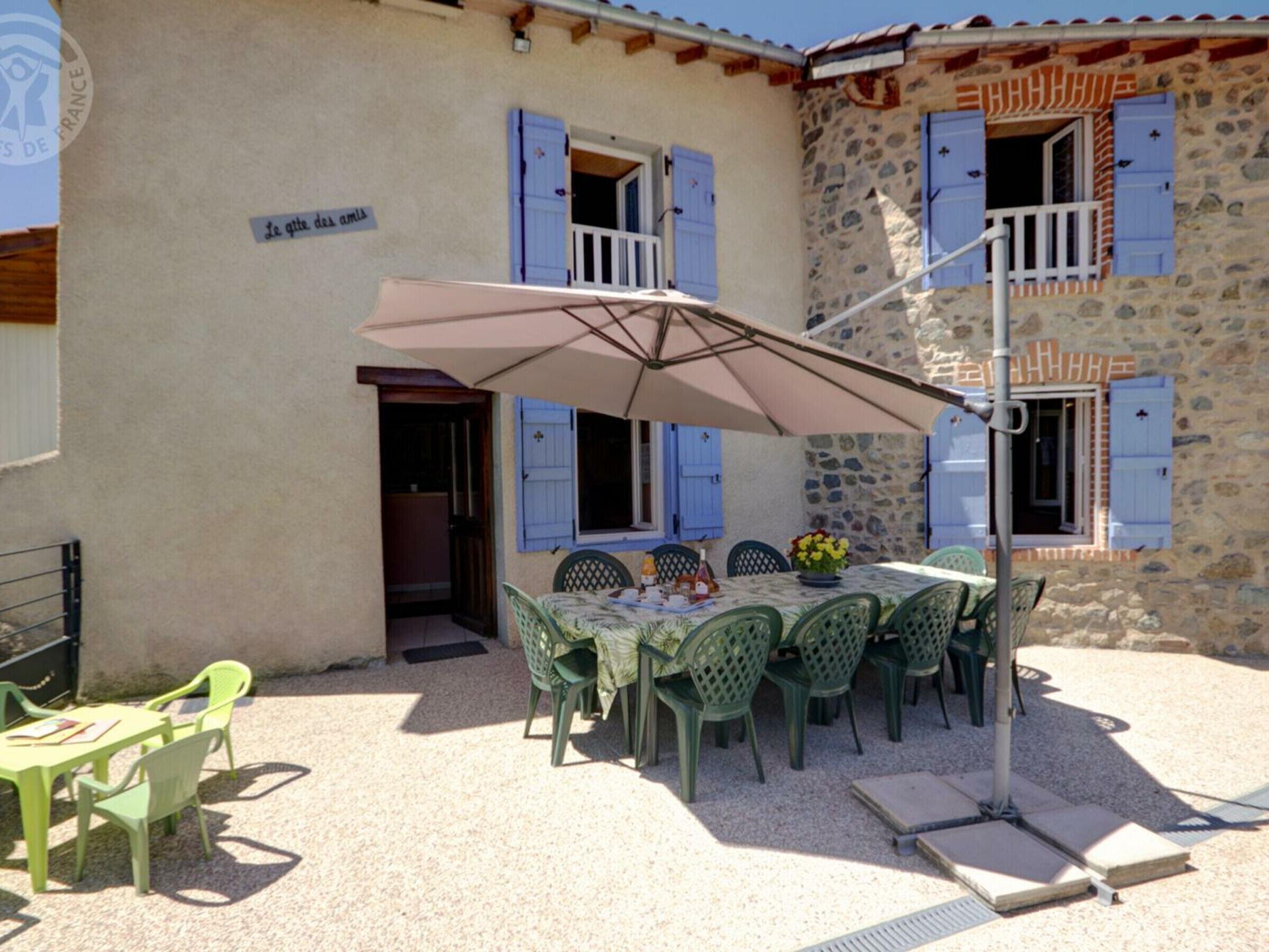 An outdoor dining area with a table and chairs, a large umbrella, and a stone building with blue shutters.