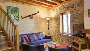 Un accogliente interno di una baita con una scala in legno, un divano con cuscini, una parete in pietra e un televisore su un supporto in legno.