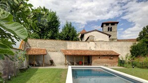 A swimming pool with a tiled deck, surrounded by a grassy area and a brick wall with a bell tower.