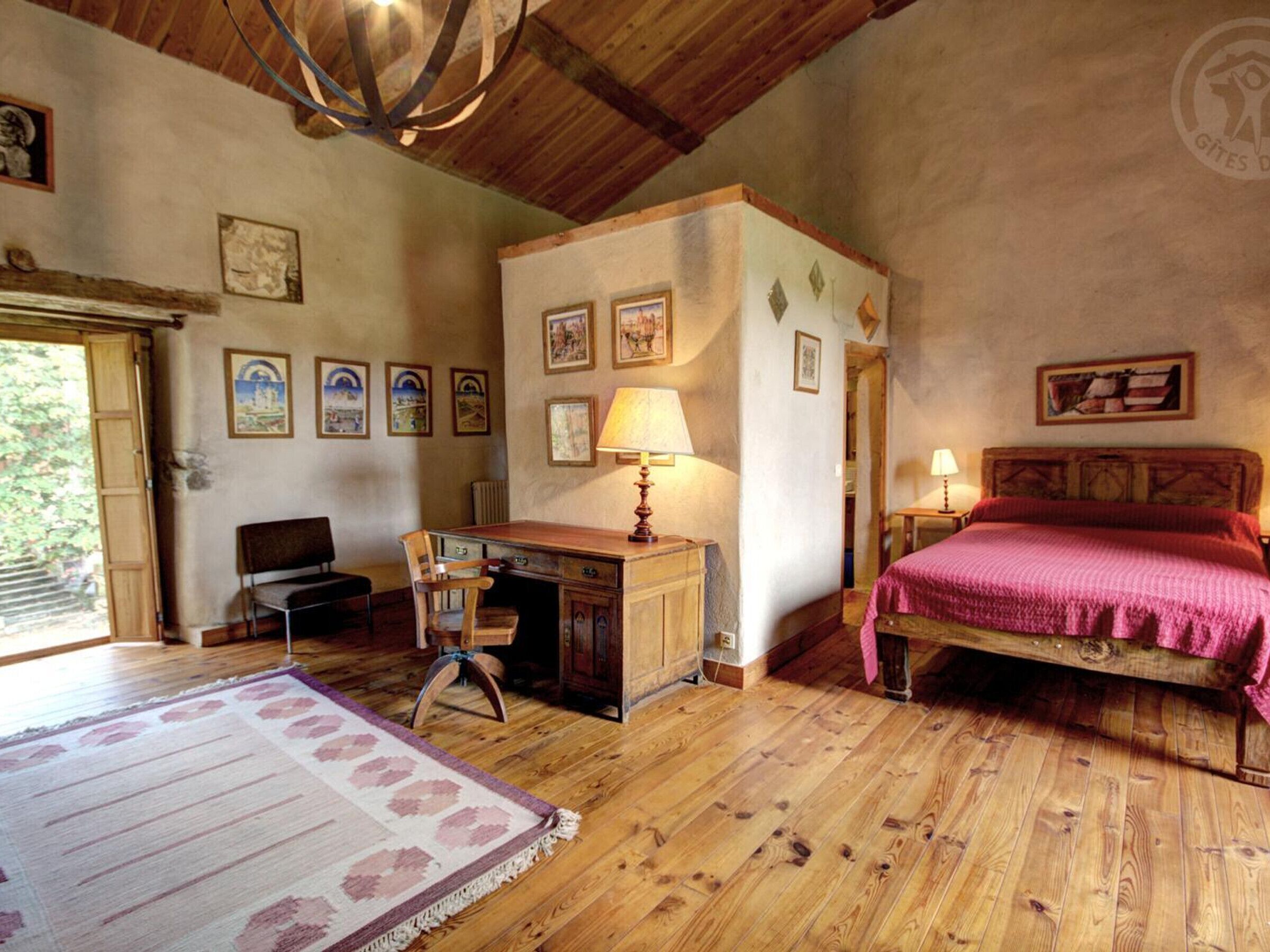 A rustic bedroom with wooden floors, a desk, a chair, a bed with a red blanket, and several framed pictures on the walls.