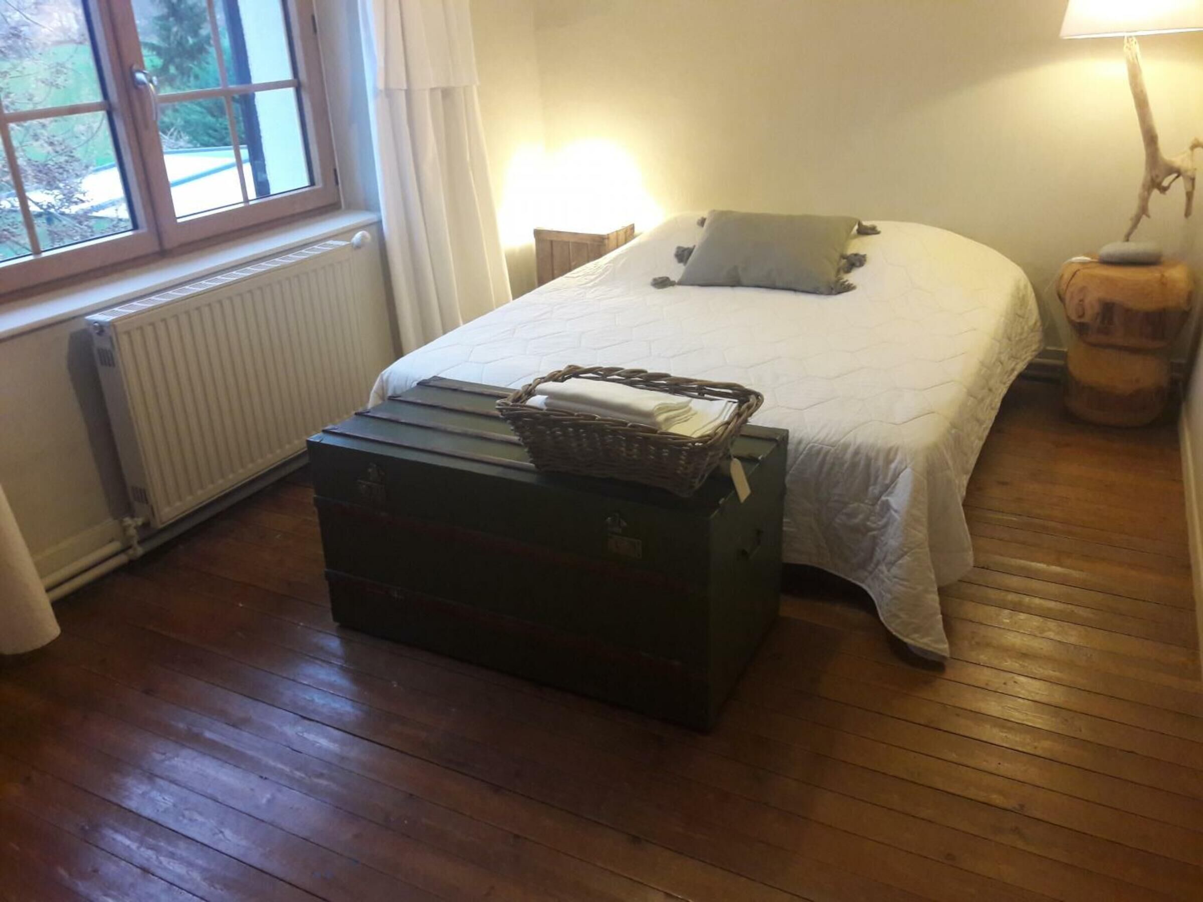 Ein Schlafzimmer mit einem Bett, einem Nachttisch, einer Lampe, einem Heizkörper, einem Fenster und einem Koffer.