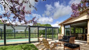 A modern outdoor patio with a glass enclosure, wooden furniture, and a view of a swimming pool. - Large house with heated private pool and pets allowed (Vivans)