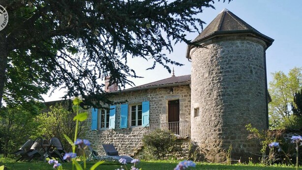 A stone house with a tower, blue shutters, and a garden with flowers.