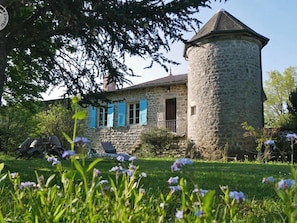 Una casa de piedra con torre, contraventanas azules y un jardín lleno de flores.