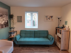 Een woonkamer met een blauwgroene bank, een witte fauteuil, een houten kast en een raam met uitzicht op een bakstenen gebouw.