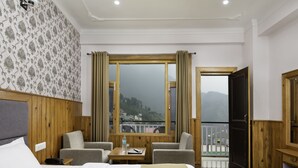 A hotel room with a bed, two armchairs, a small table with a bottle and a remote control, a window with curtains, and a view of a mountainous landscape. - Hotel Mount View (Dharamshala)