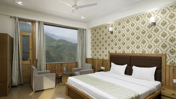 A hotel room with a large bed, a desk, a chair, a small table, and a view of mountains through a large window. - Hotel Mount View (Dharamshala)