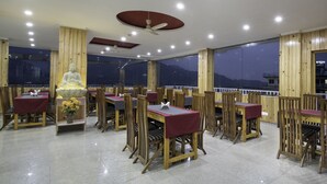 A restaurant with wooden furniture, a Buddha statue, and large windows offering a view of the mountains. - Hotel Mount View (Dharamshala)