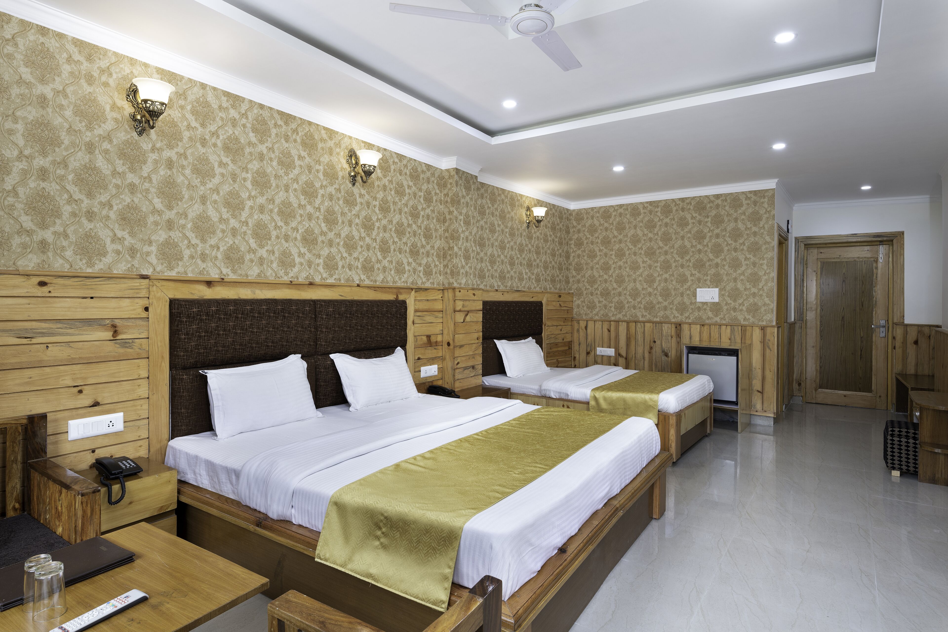 A hotel room with two beds, wooden headboards, a ceiling fan, and a small desk with a phone and a remote control.
