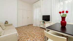 A room with a sofa, a TV on a stand, a dining table with a vase of red flowers, and a tiled floor. - Rosso apartment, first floor (Unknown)