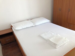 A neatly made bed with white linens and pillows, a wooden nightstand with a drawer, and a wooden wardrobe with two drawers. - The White house robinson (Gdinj)