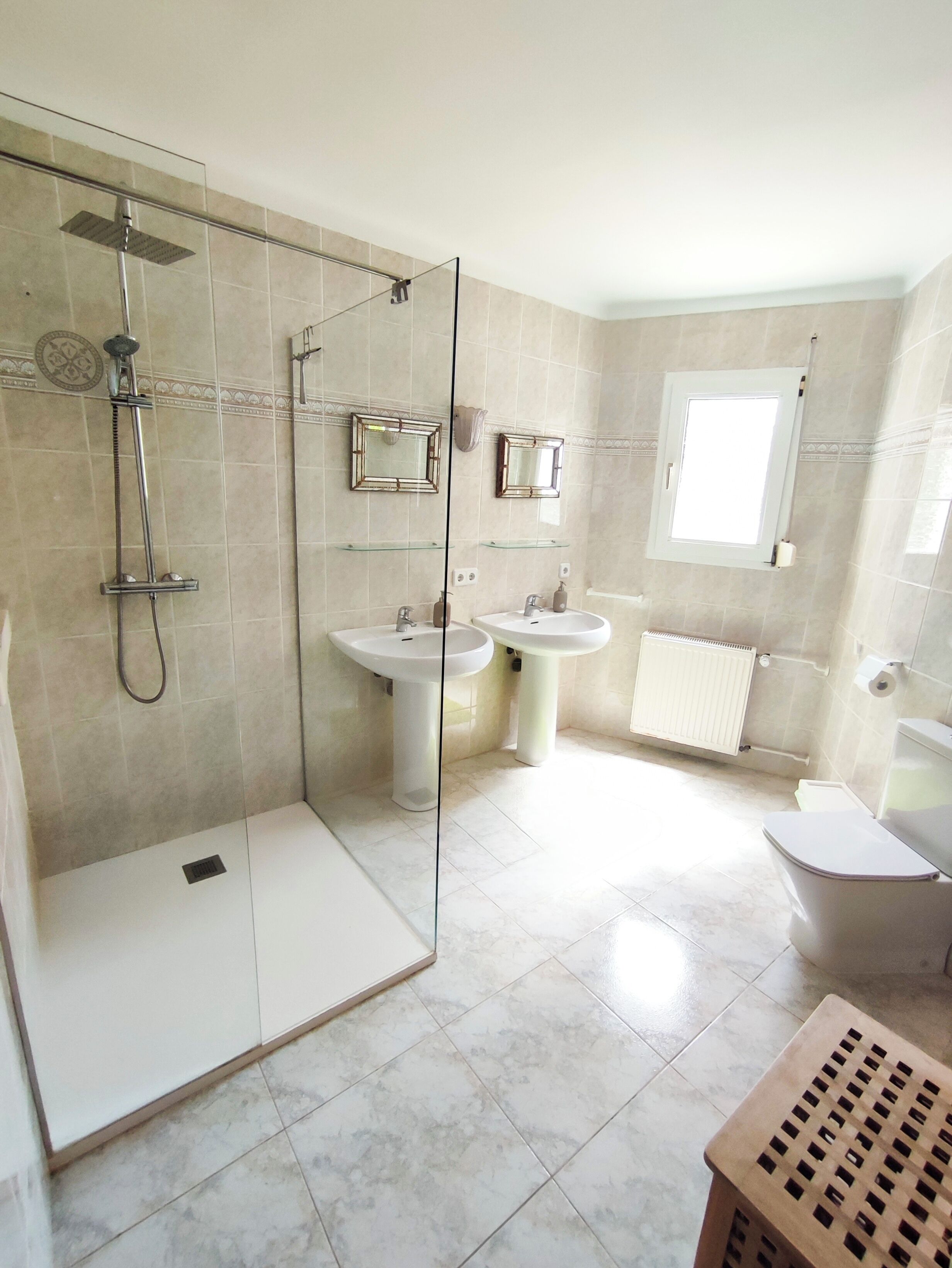 A modern bathroom with a glass-enclosed shower, two sinks, a toilet, and a window.