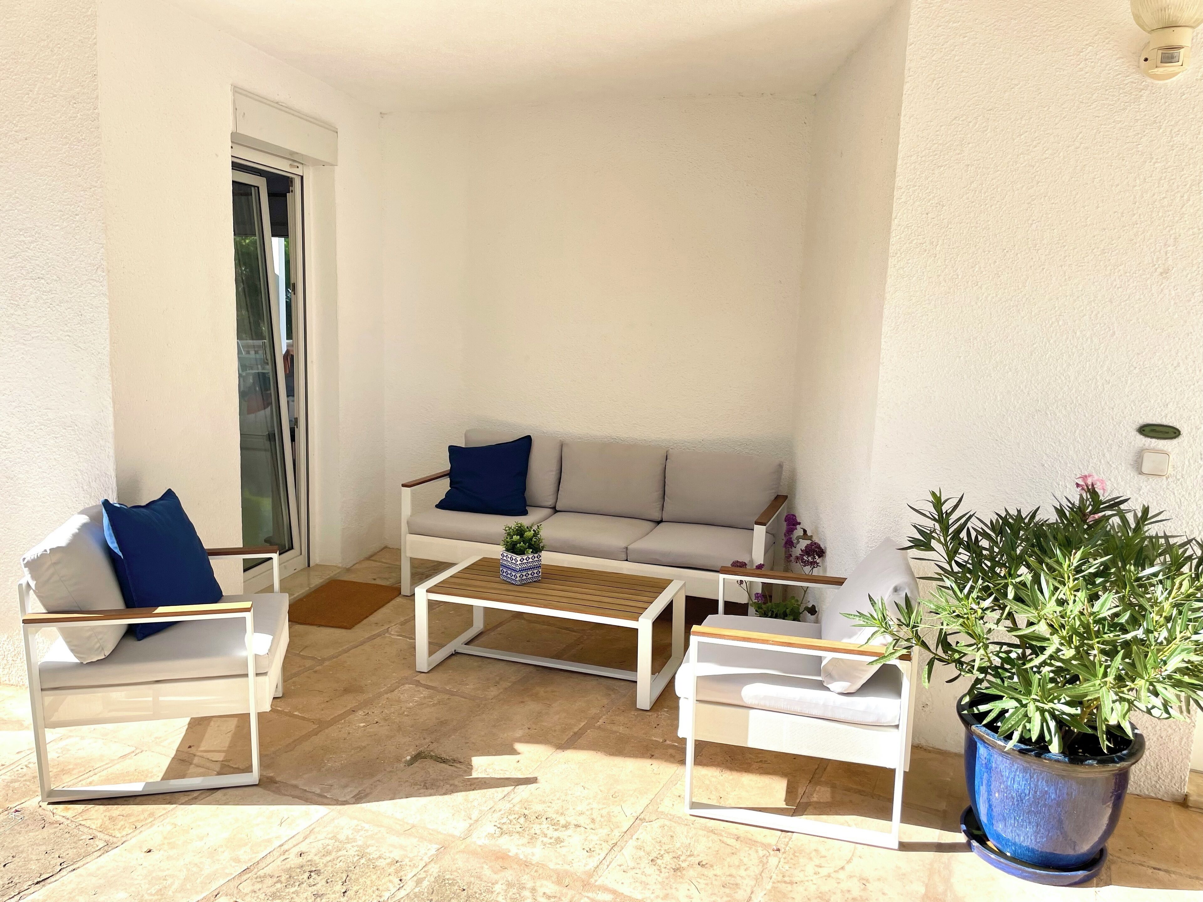 A sunlit patio with a white sofa, two white chairs, a wooden coffee table, and a potted plant.