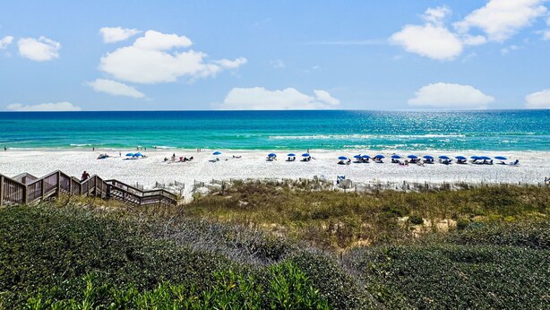 Een strand met een houten promenade die naar het strand leidt, helder turquoise water en een zandstrand met parasols.