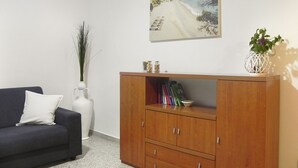 Una sala de estar con un sofá oscuro, un jarrón blanco, un mueble de madera y un cuadro con una playa.