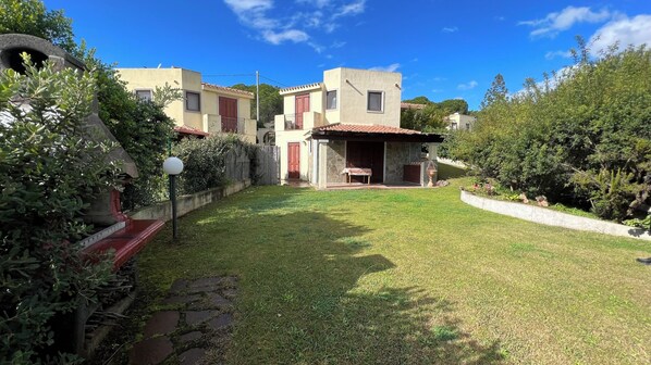 A residential area with a stone archway, a small patio with a table and chairs, and a grassy yard. - Chia: Cottage with garden, air conditioning, barbecue,and parking. (Eden Rock)