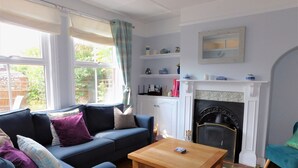 A living room with a blue sofa, a wooden coffee table, a fireplace, and a view of the garden through large windows. - 1 Orchard Cottages -  a cottage that sleeps 5 guests  in 2 bedrooms (Cromer)