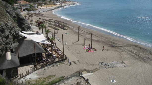 Una playa con un pasillo de madera, sombrillas y sillas. Se ven edificios al fondo y un cielo despejado.