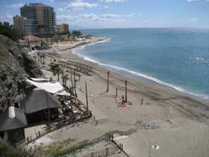 A beach with a wooden walkway, umbrellas, and beach chairs. There are buildings in the background and a clear sky. - Torreon del Mar BeachFront apartment (Benalmádena)