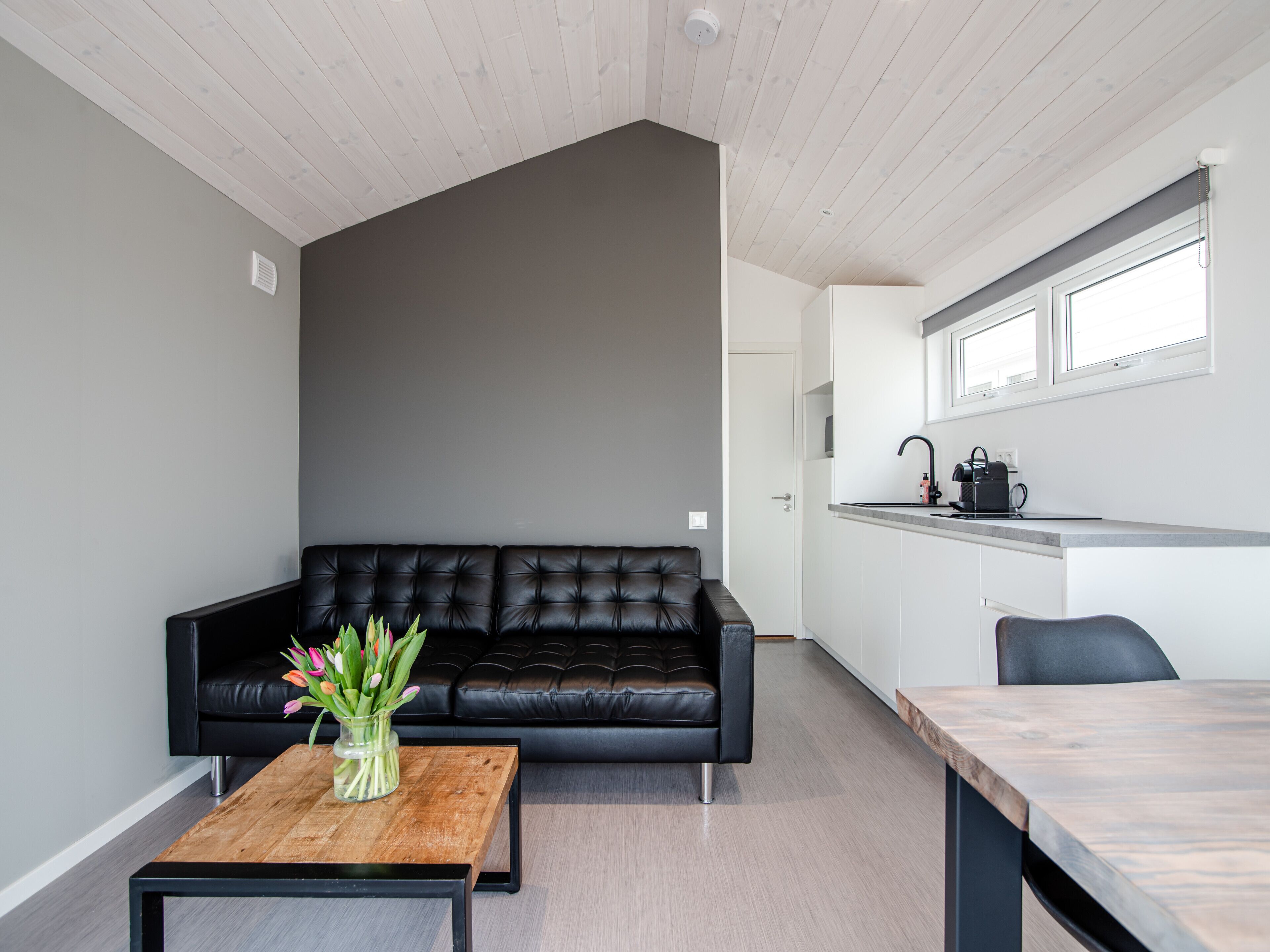 A modern living room with a black leather sofa, a wooden coffee table with a vase of flowers, and a kitchen area with a countertop and sink.