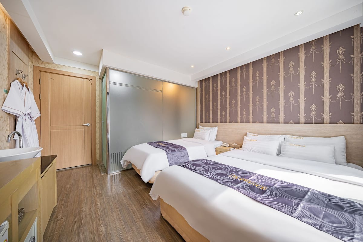 A hotel room with two beds, a wooden wardrobe, a bathroom with a sink, and a patterned wallpaper on the wall.