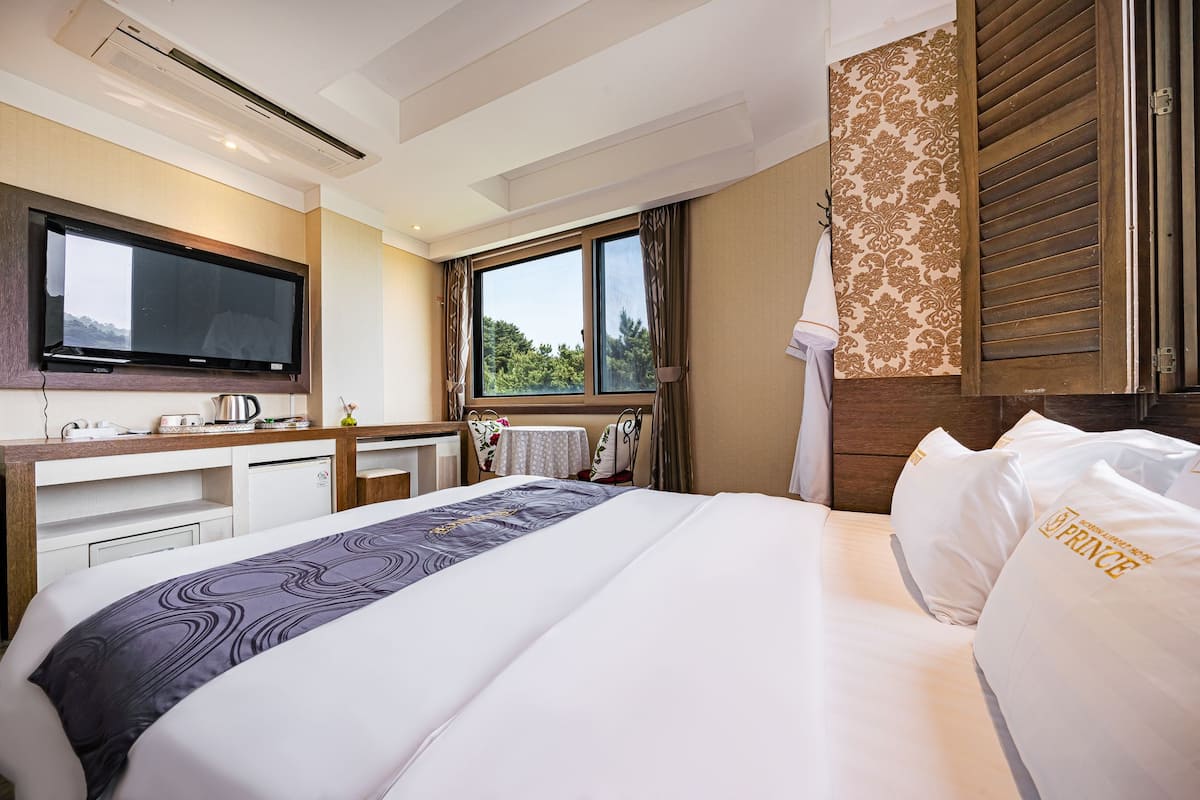 A hotel room with a bed, a desk with a flat-screen TV, a window with curtains, and a wall-mounted air conditioner.