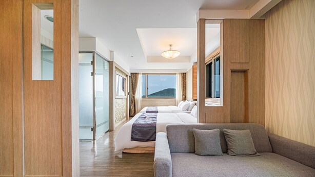 A hotel room with a wooden floor, a gray sofa, a bed with white and purple bedding, and a view of a mountain through the window. - Incheon Prince Tourist Hotel (Incheon)