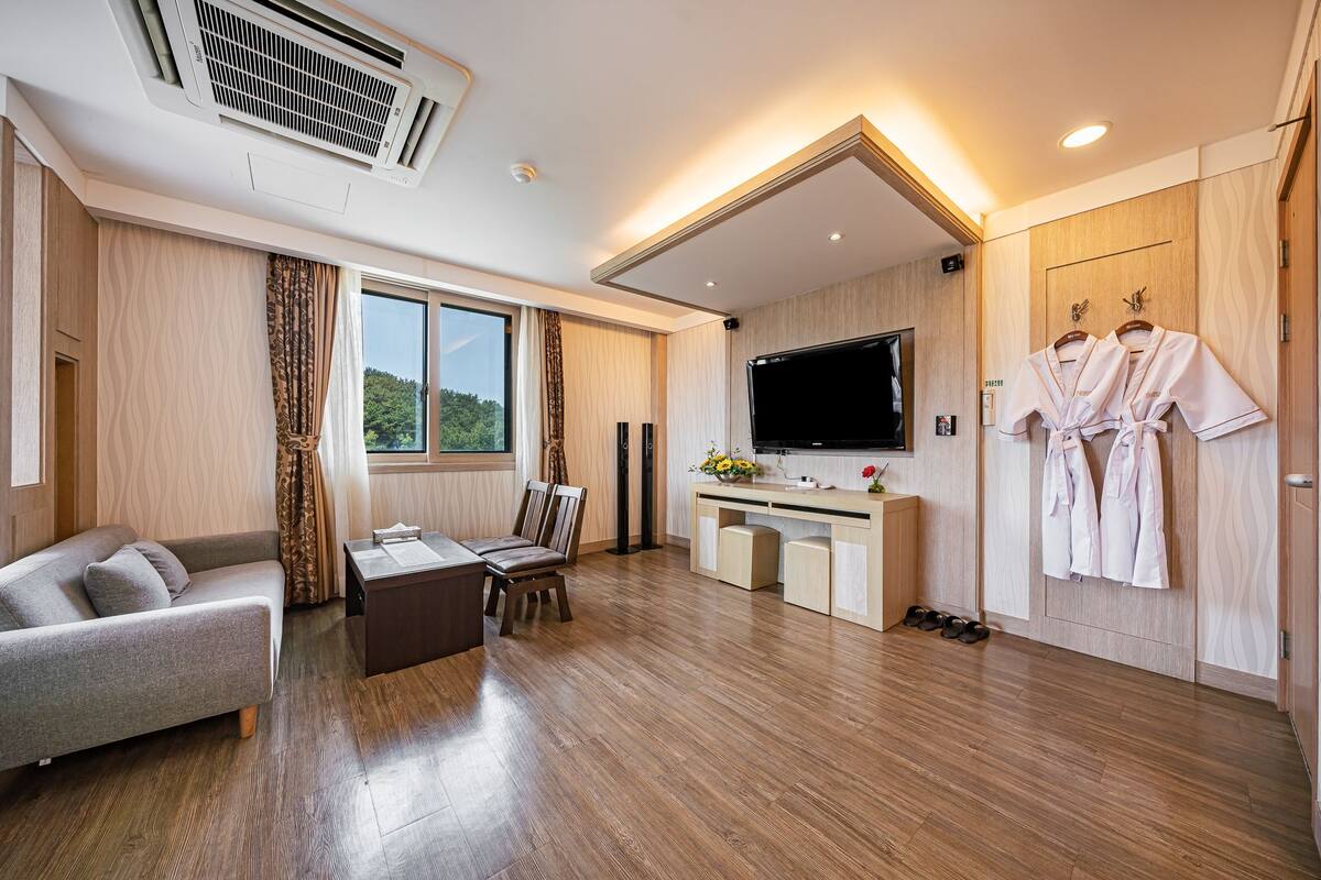 A modern hotel room with a sofa, dining table, chairs, a TV, a desk with a computer, and a wardrobe with robes.