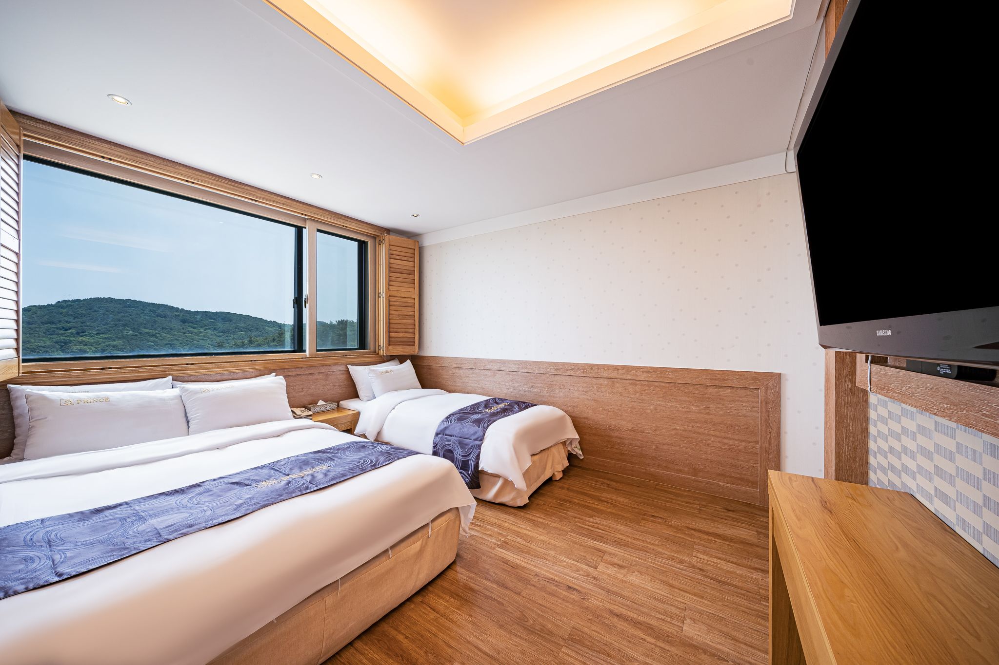 A hotel room with two beds, a large window, a flat-screen TV, and a wooden floor.