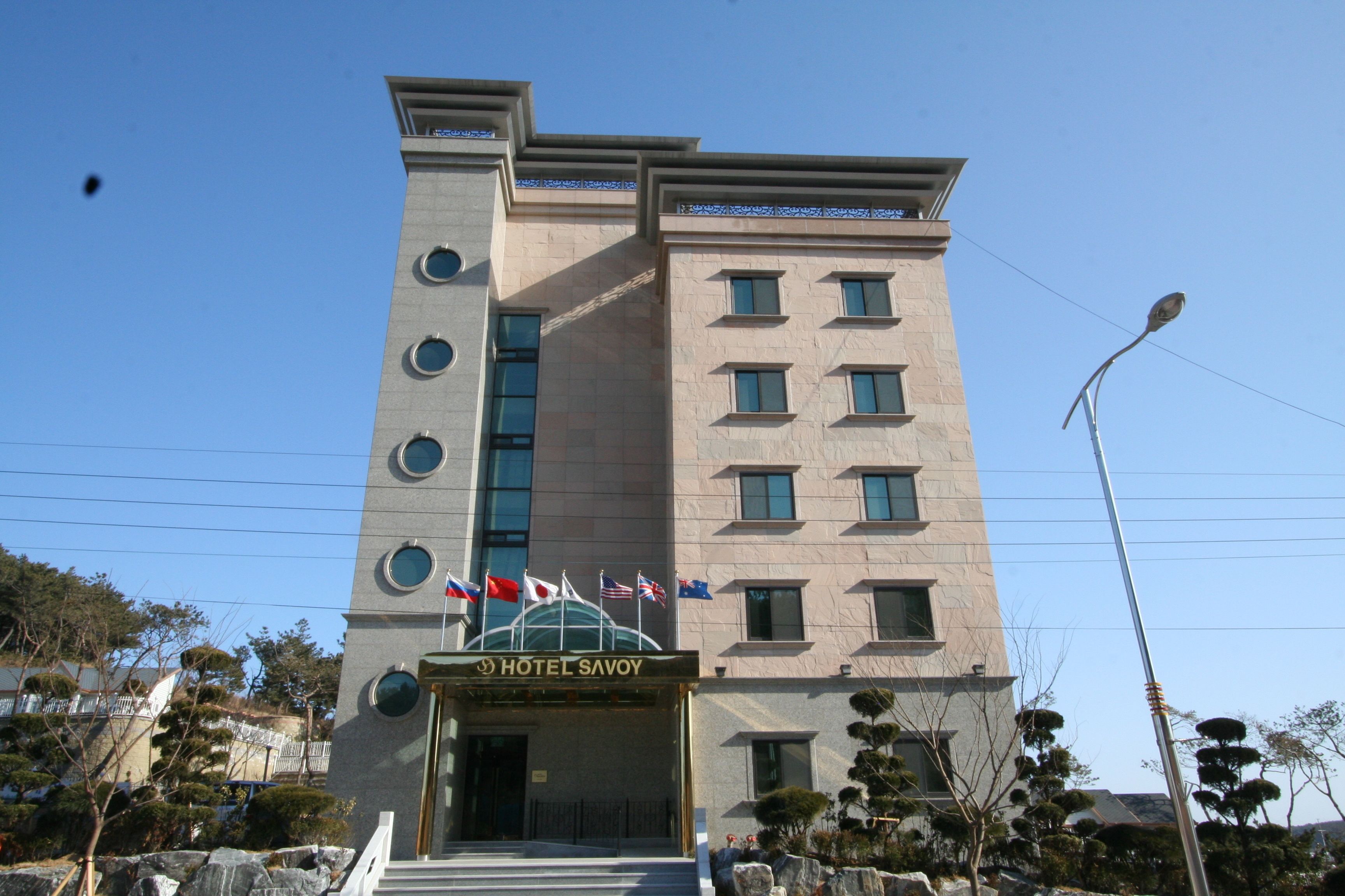 A multi-story hotel with a modern design, featuring a prominent entrance with flags, a series of windows, and a clear sky in the background.