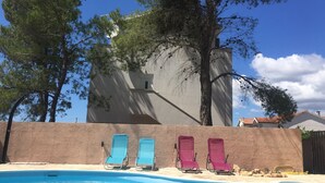 Une piscine avec des chaises longues, un bâtiment doté d’un mur à motifs, et des arbres en arrière-plan.