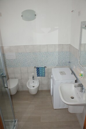 Une salle de bain comprenant des toilettes, un bidet, un lave-linge, un lavabo et une douche à parois de verre.